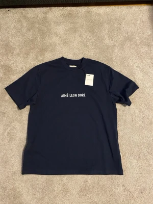 Mörkblå t-shirt från Aimé Leon Dore - Snygg mörkblå t-shirt från Aimé Leon Dore med vit logotyp på bröstet. Klassisk passform med rund hals och korta ärmar. Tillverkad i mjuk bomull som känns skön mot huden. Perfekt för en clean och stilren look.