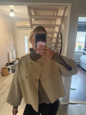 Kort beige cape-rock med guldknappar - Stilren beige cape-rock med breda ärmar och dubbelknäppning med stora guldknappar. Rocken har en boxig, kort passform och är tillverkad i ett tjockare tyg. Finns små fläckar men inget jag ser med blotta ögon (inte testat tvätta). Finns inte att köpa längre 💕