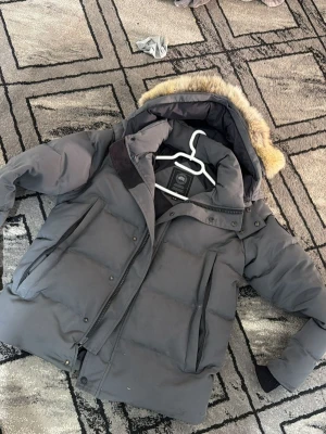 Grå dunjacka från Canada Goose - Snygg grå dunjacka från Canada Goose med stor huva och päls. Jackan har dragkedja, tryckknappar och två stora fickor framtill. Perfekt för kalla vinterdagar tack vare det tjocka dunfodret och vindtäta materialet.