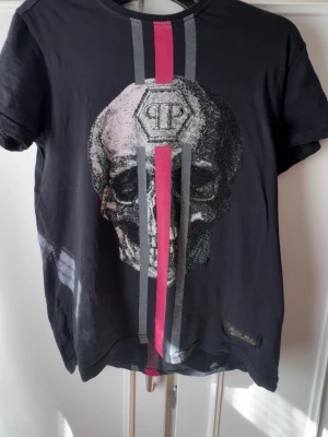 Svart t-shirt med dödskalle från Philipp Plein - Cool svart t-shirt från Philipp Plein med stort dödskalletryck framtill och röda samt grå ränder både fram och bak. T-shirten har rund hals och korta ärmar. Märkesdetalj nertill och PP-logga på ryggen. Perfekt för dig som gillar edgy streetstyle.