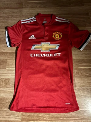 Manchester United matchtröja Adidas - Röd Manchester United matchtröja från Adidas med klubbmärke på bröstet, vita Adidas-ränder på axlarna och Chevrolet-logga framtill. Tröjan har korta ärmar med svart och vit kant, samt knappslå vid halsen. Tillverkad i lätt funktionsmaterial.