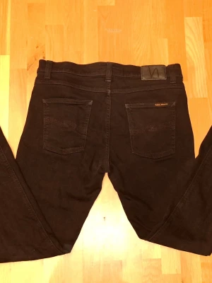 Svarta jeans från Nudie Jeans - St: W33 L36 Snygga svarta jeans från Nudie Jeans med klassisk femficksmodell och raka ben. Jeansen har en diskret logga på bakfickan och patch i läderimitation vid linningen. Tillverkade i mjukt denimtyg som sitter skönt hela dagen.
