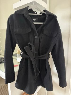 Svart ullkappa från Bik Bok - Stilren svart kappa från Bik Bok i ullblandning. Modellen har knappar framtill, två bröstfickor med lock och ett knytbälte i midjan för en snygg siluett. 