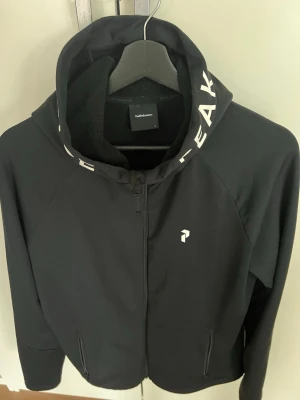 Svart hoodie från Peak Performance - Snygg svart hoodie från Peak Performance med dragkedja framtill och vit logga på bröstet. Huva med vit textdetalj längs kanten och två fickor med dragkedja. Perfekt för en sportig och avslappnad stil. Varm och skön