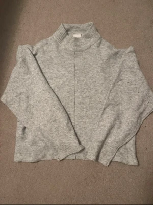 Ljusgrå stickad polotröja - Mysig ljusgrå stickad tröja med hög krage och lång ärm. Tröjan har en rak passform, ribbade muddar och en dekorativ söm framtill. Perfekt för kyliga dagar och enkel att matcha till olika outfits.