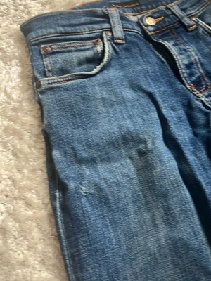  Blåa Nudie jeans  - Säljer ett par klassiska blå jeans i slim modell med slitningar och hål vid benslutet. Jeansen har fem fickor och kontrastsömmar. Perfekta för en stilren ”grisch” look