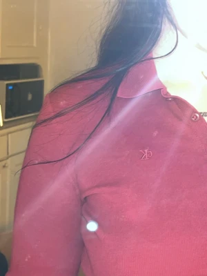 Röd långärmad topp från Calvin Klein - Säljer en röd långärmad topp från Calvin Klein med klassisk pikékrage och knappar vid halsen. Toppen är croppad och har en liten CK-logga broderad på bröstet. Perfekt till jeans eller kjol för en clean och snygg look.