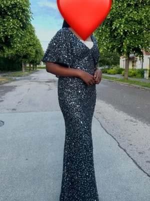 Glittrig långklänning med v-ringning - Säljer en elegant långklänning i mörkblått med glittriga paljetter över hela klänningen. Klänningen har v-ringning, korta vida ärmar och figurnära passform som framhäver siluetten. Perfekt för bal eller festliga tillfällen.