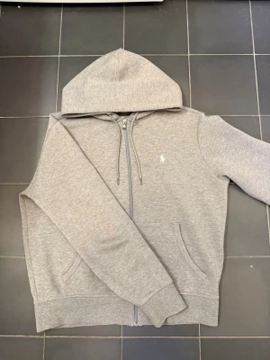 Grå hoodie från Polo Ralph Lauren - Snygg grå hoodie från Polo Ralph Lauren med dragkedja, huva med dragsko och två fickor framtill. Klassisk logga broderad på bröstet. Tillverkad i mjukt sweatshirtmaterial som är riktigt skönt att ha på sig.