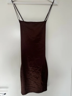 Brun satin klänning med tunna band - Snygg brun kortklänning från H&M i satin med tunna axelband och öppen rygg med snygga banddetaljer. Klänningen har en figurnära passform och är perfekt för dig som gillar minimalistisk och trendig stil.