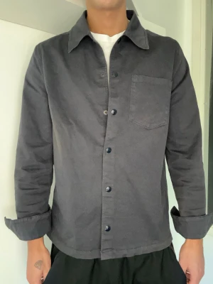 Mörkgrå overshirt med bröstficka - Stilren mörkgrå overshirt med klassisk krage och knäppning framtill. Overshirten har en bröstficka på vänster sida och raka ärmar med knapp vid ärmslut. Tillverkad i ett mjukt bomullstyg som ger en avslappnad look.