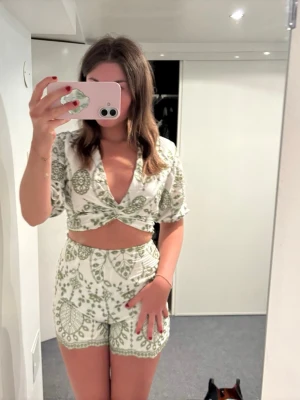 Grönt och vitt set med mönster - Säljer ett snyggt tvådelat set i vitt med grönt blommigt och paisley-inspirerat mönster. Toppen har omlott och djup urringning samt korta puffärmar. Shortsen är höga i midjan och har matchande mönster. Perfekt för dig som vill sticka ut lite extra!