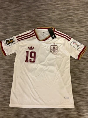 Vit Spanien fotbollströja Adidas - Säljer en vit Spanien fotbollströja från Adidas med röda detaljer, nummer ”Lamine Yamal 19” och officiella märken på ärmarna. Tröjan har guldrandig krage och röda ränder på axlarna. Tillverkad i lätt och ventilerande material, perfekt för match eller träning.🇪🇸1️⃣9️⃣