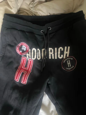 Svarta Hoodrich mjukisbyxor med patchar - Säljer ett par svarta mjukisbyxor från Hoodrich med flera snygga patchar och broderade detaljer i vitt och rött. Byxorna har dragsko i midjan och fickor på sidorna. Perfekta för en streetwear-look och riktigt bekväma.