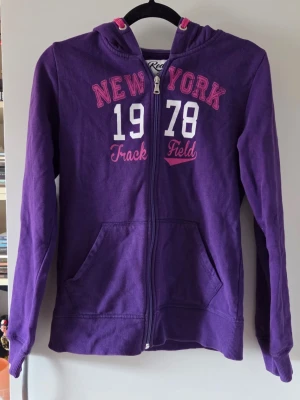 Lila hoodie med dragkedja och tryck - Lila hoodie med huva och dragkedja framtill. Tryck på bröstet med texten 'NEW YORK 1978 Track Field' i vitt och rosa. Kängurufickor och ribbade muddar vid ärmslut och nederkant. Mjuk insida och avslappnad passform, perfekt för chill dagar. Från barnavdelningen så storleken motsvarar ungefär xs/s.