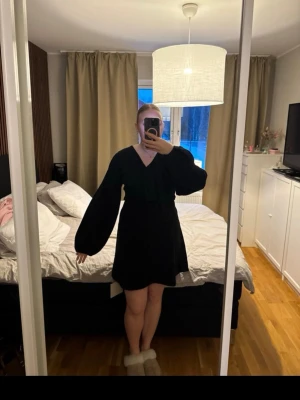 Svart omlottklänning med vida ärmar - Kort svart omlottklänning med v-ringning och vida långärmar. Klänningen har en croppad effekt i midjan som kan justeras för mer eller mindre öppen look. Stilren och modern design som ger en edgy vibe.
