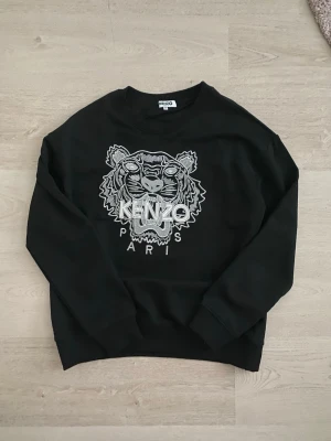 Svart sweatshirt från Kenzo - Svart sweatshirt från Kenzo med stort broderat tigerhuvud och texten 'KENZO PARIS' på bröstet. Tröjan har rund halsringning och ribbade muddar vid ärmslut och nederkant. Perfekt för dig som gillar streetwear och vill sticka ut.
