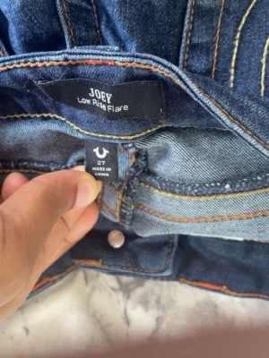 Mörk blå true religion jeans - Säljer ett par blå jeans med låg midja och utsvängda ben från Joey. Jeansen har dekorativa sömmar och fickor bak med detaljer. Perfekta för dig som gillar en retroinspirerad look och vill ha ett par riktigt snygga flare jeans.