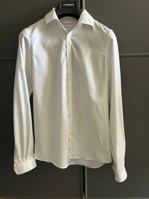 Vit strykfri skjorta från Lindbergh - Klassisk vit slim fit skjorta från Lindbergh med lång ärm och knappar framtill. Skjortan har en tidlös design med krage och är tillverkad i ett mjukt bomullsmaterial. Perfekt för en clean och stilren look. Skjortan har dessutom nanotex-skydd mot fläckar samt antibakterial behandling som skyddar mot lukt och bakterier. Nypris 999kr