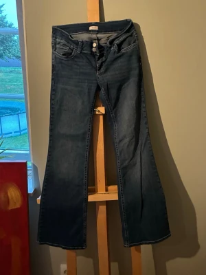 Mörkblå bootcut jeans från Nelly - Snygga mörkblå bootcut jeans från Nelly med deras ikoniska fickor. Jeansen har en lätt utsvängd passform nedtill och är tillverkade i denim med en skön stretch. Självfixat håll vid en av banden på midjan, inte så synligt när jeansen är väl på. Priset går att diskuteras. Perfekta för dig som gillar en retroinspirerad look.