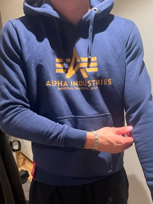 Blå hoodie Alpha Industries - Snygg blå hoodie från Alpha Industries med guldfärgat tryck på bröstet och klassisk känguruficka. Huva med dragsko och ribbade muddar vid ärmar och midja. Perfekt för en avslappnad streetwear-look.