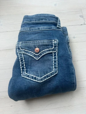 Blå bootcut jeans med slitningar - Säljer ett par blå bootcut jeans med slitningar på benen och snygga kontrastsömmar på bakfickan. Jeansen har låg midja och klassisk femficksdesign. Jeansen är i bra skick och är storlek 170.