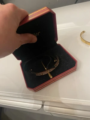 Guldigt armband med stenar - Snyggt guldigt armband med gnistrande stenar och dekorativa detaljer. Kommer i en röd ask med gulddetaljer. Armbandet har en klassisk form och är prytt med flera små stenar som ger en lyxig känsla.
