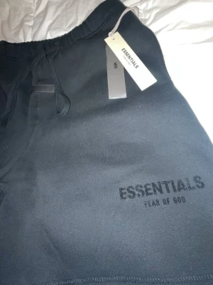 Svarta shorts från Essentials Fear of God - Svarta shorts från Essentials Fear of God med diskret logga framtill. Modellen har dragsko i midjan för justerbar passform och är tillverkad i mjukt bomullsmaterial. Perfekta för en avslappnad och trendig look.