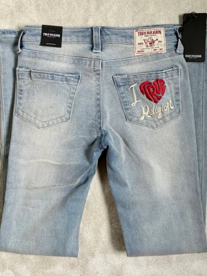 Ljusblå True Religion jeans - Säljer ett par ljusblå jeans från True Religion med broderad bakficka där det står 'I True Religion' i vitt och rött. Jeansen har klassisk femficksmodell och raka ben. Materialet är mjukt denim och de har en snygg tvättad look.