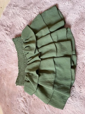 Grön volangkjol med smock - Supersöt grön kortkjol med bred smock i midjan och flera lager volanger. Kjolen har en luftig känsla och passar perfekt till varma dagar. Enkel att matcha med både toppar och sneakers för en chill look.