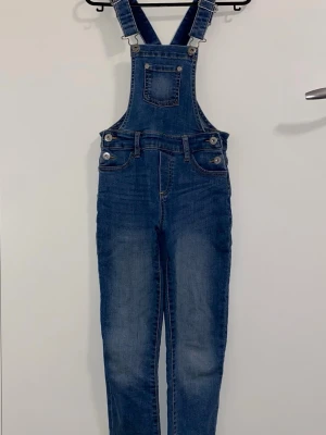 Blå denim byxdress med hängslen - Säljer en klassisk blå jeansbyxdress med justerbara hängslen och fickor fram. Byxdressen har smala ben och silverfärgade knappar i sidorna samt på hängslena. Perfekt för en avslappnad och trendig look.