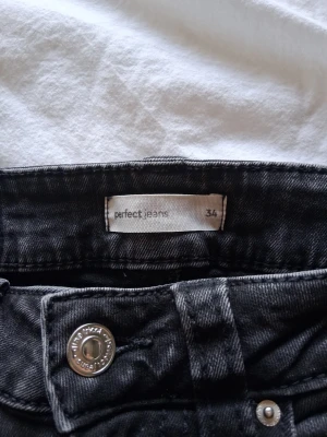 Svarta bootcut jeans, storlek 34 - Säljer ett par svarta bootcut jeans med klassisk femficksdesign och silverfärgade knappar. Jeansen har en lätt tvättad look och är tillverkade i ett mjukt denimtyg som sitter snyggt på benen. Perfekta till en avslappnad stil.