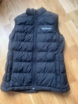 Svart dunväst Peak Performance XS - Snygg svart dunväst från Peak Performance i storlek XS. Västen har hög krage, dragkedja framtill och diskret logga på bröstet. Perfekt för lager-på-lager och håller dig varm med sitt fluffiga dunmaterial.
