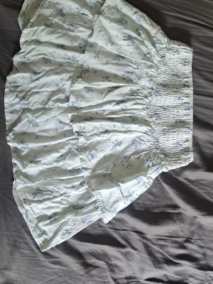 Hollister vit blommig kjol - Söt kort kjol från Hollisteri vitt med ljusblått blommönster. Kjolen har volanger och är gjord i ett mjukt bomullstyg som känns luftigt och perfekt för varma dagar.