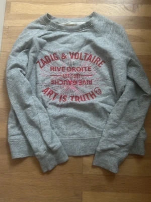 Grå sweatshirt Zadig & Voltaire - Snygg grå sweatshirt från Zadig & Voltaire med röd text och tryck framtill där det står 'ART IS TRUTH'. Tröjan har rund halsringning, långa ärmar och ribbade muddar. Perfekt för en avslappnad och trendig look.