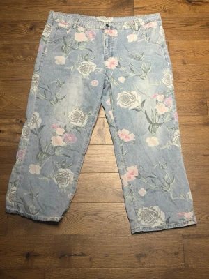 Blommiga ljusblå jeans med raka ben - Unika ljusblå jeans med stort blommönster i vitt, rosa och grönt. Jeansen har raka ben, klassisk femficksmodell och normal midja. Perfekta för dig som vill sticka ut med en cool och trendig look. Materialet är klassisk denim i bomull.