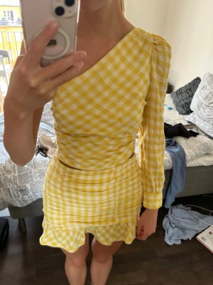 Gult sett - Säljer ett gulligt sett i gul och vit rutig design. Toppen har oneshoulder-modell med en lång ärm och kjolen har volangkant nertill. Materialet känns lätt och passar perfekt till varmare dagar. Figurnära passform och trendig look. Den är i M men är liten i storleken, passar mig som brukar ha S.