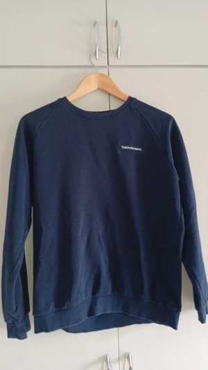 Sweatshirt Peak Performance - En stilren mörkblå sweatshirt från Peak Performance med rund halsringning och diskret logga på bröstet. Tröjan har långa ärmar, ribbade muddar och är tillverkad i mjukt bomullsmaterial. Perfekt för en avslappnad och snygg look.