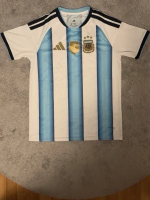 Argentina matchtröja Adidas blåvit - Säljer en officiell Argentina matchtröja från Adidas med klassiska blå och vita ränder, svarta detaljer och guldiga emblem på bröstet. Tröjan har korta ärmar, rund hals och är tillverkad i lätt funktionsmaterial. Perfekt för fotbollsfans!