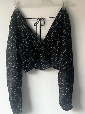 Svart  prickig blus med knyt - Trendig svart blus med vita prickar och djup v-ringning. Toppen har långa ärmar och knytband i ryggen för en snygg detalj. 