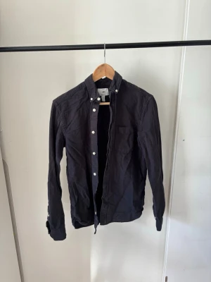 Mörkblå skjorta med knappar - Stilren mörkblå skjorta med vita knappar och button-down krage. Skjortan har lång ärm och bröstficka på vänster sida. Perfekt till jeans eller chinos för en clean look.