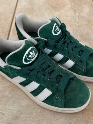 Mörkgröna Adidas Campus 00s - Snygga gröna Adidas Campus 00s med klassiska vita ränder på sidorna och vit sula. Skorna har gröna skosnören och en tydlig Adidas-logga på plösen. Låg modell som passar perfekt till streetwear och casual outfits. Kolla etiketten för närmare preciseringar av storleken. 