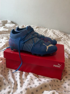 Blåa Puma Future fotbollsskor - Blåa Puma Future fotbollsskor med vita och röda detaljer. Skorna har snörning, låg profil och en mönstrad ovandel för extra grepp. Stor vit Puma-logga på sidan och texten FUTURE i vitt. Designade för spel på både konstgräs och gräs.