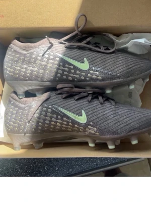 Nike Vapor 16 Elite KM AG-Pro - Säljer ett par Nike Vapor 16 Elite KM AG-Pro fotbollsskor i färgen plum eclipse med metallic silver detaljer och ljusgrön Swoosh. Skorna har låg profil, snörning och dobbar för konstgräs. Materialet är syntetiskt och designen är modern och sportig.