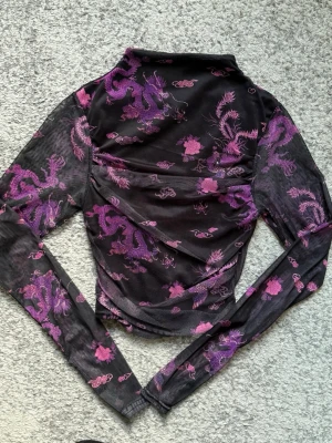 Svart mesh topp med lila drakmönster - Snygg svart mesh topp med lila drakmönster och blommor. Toppen är långärmad och har en tight passform med hög hals. Perfekt för dig som vill sticka ut med ett unikt asiatiskt inspirerat print.