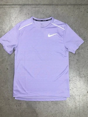 Nike träningströja - Snygg ljuslila tränings-tshirt från Nike med klassisk logga på bröstet och detaljer vid axlarna. Tillverkad i ett lätt och snabbtorkande material som passar perfekt till gymmet eller löprundan. Kort ärm och rund hals.