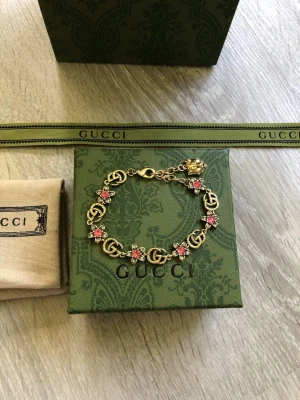 Gucci armband med rosa detaljer - Snyggt armband från Gucci i guldfärgad metall med ikoniska GG-loggor och små rosa blommor mellan länkarna. Armbandet har ett justerbart lås och kommer med originalförpackning, dustbag och presentpåse. Perfekt accessoar för att lyfta din stil.