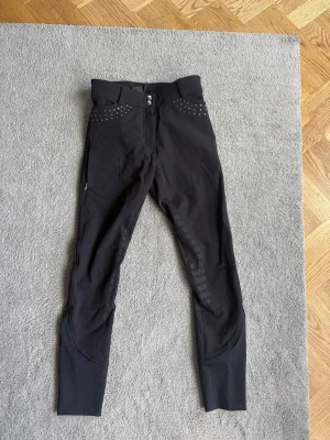 Svarta ridbyxor CRW - Snygga svarta ridbyxor med hög midja och silikongrepp på insidan av benen för extra fäste. Dekorativa nitar vid fickorna och breda benslut i stretchigt material. Perfekta för träning eller tävling inom ridsport. Ny utan prislapp