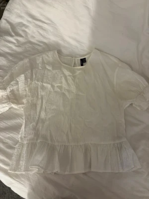 Vit volangblus med kort ärm - Söt vit blus i lätt och luftig bomull med korta ärmar och volang nedtill. Blusen har rund halsringning och en liten knapp i nacken. Perfekt till jeans eller kjol för en fräsch look.