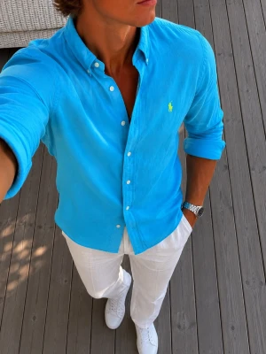 Blå skjorta från Ralph Lauren - Klassisk blå skjorta från Ralph Lauren med button-down krage och limegrönt broderat logotyp på bröstet. Skjortan har lång ärm, vita knappar och är tillverkad i bomull. Skavanker syns på bilder, några fläckar. Perfekt för en clean och stilren look.